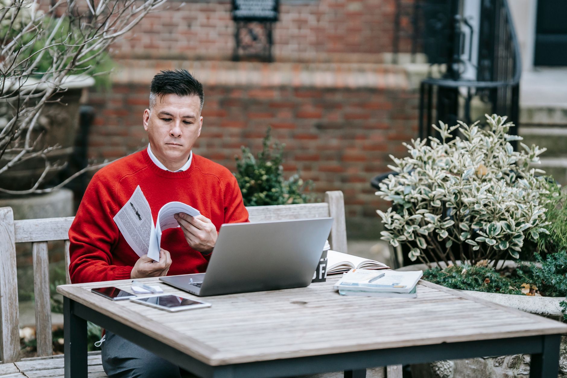 Adult male reading outside with a laptop on a wooden table, embracing remote work lifestyle.